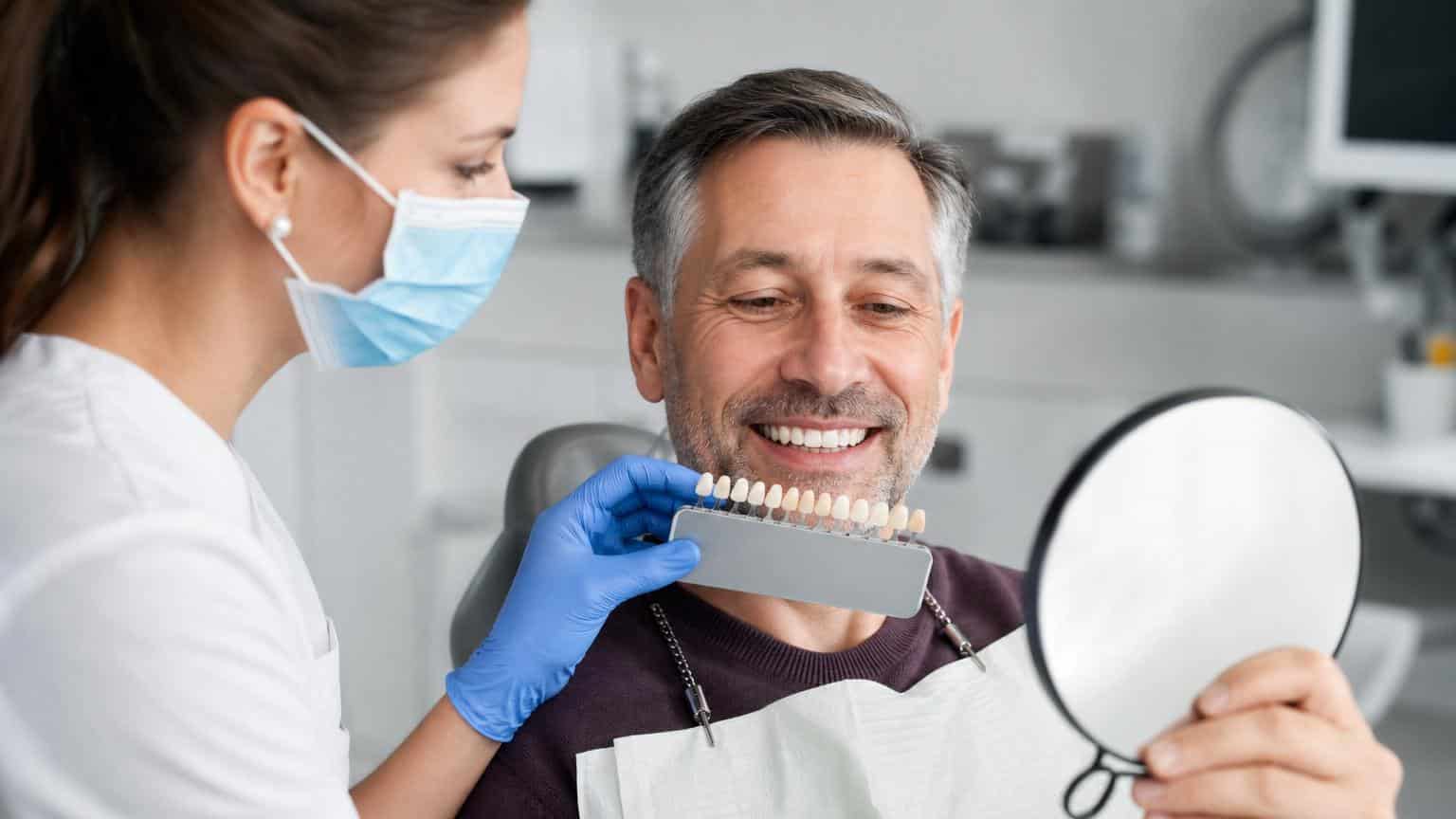 Dentist holding a tooth shade guide against a smiling man's teeth to match the color for a ceramic dental bridge.
