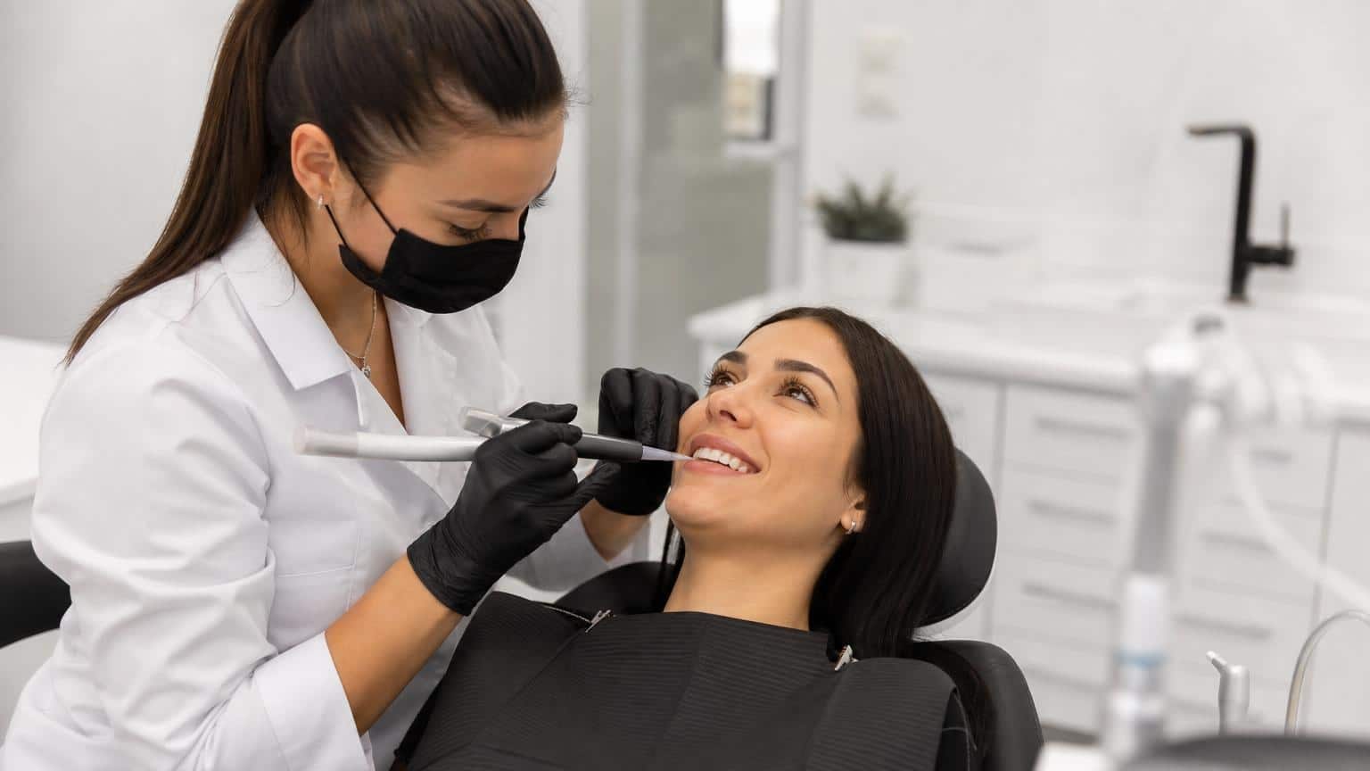 Dentist in a white coat and black mask applying composite resin to a smiling patient's teeth using a specialized injection tool.