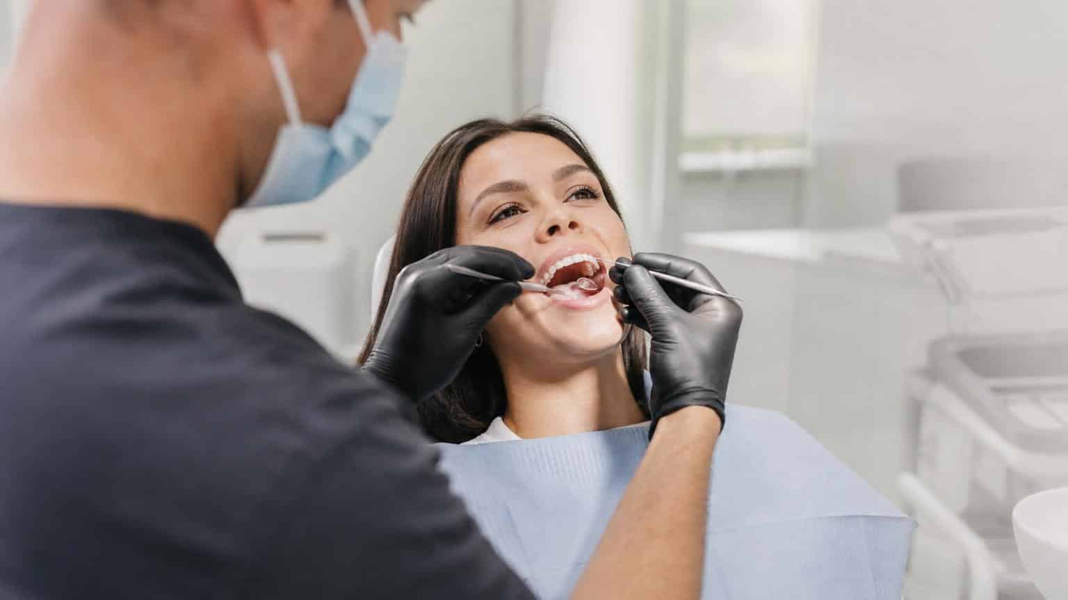 Dentist in black gloves using a mirror and probe to examine the teeth of a smiling woman in a bright, modern dental clinic.