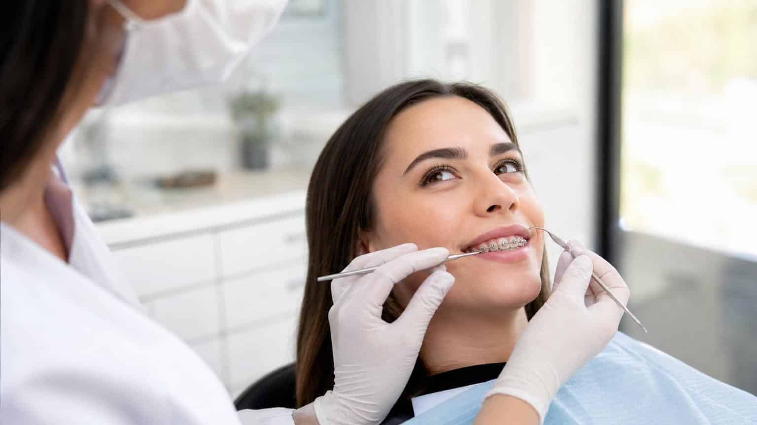 Smiling woman with metal dental braces receiving a checkup from a dentist using professional tools in a bright clinic.