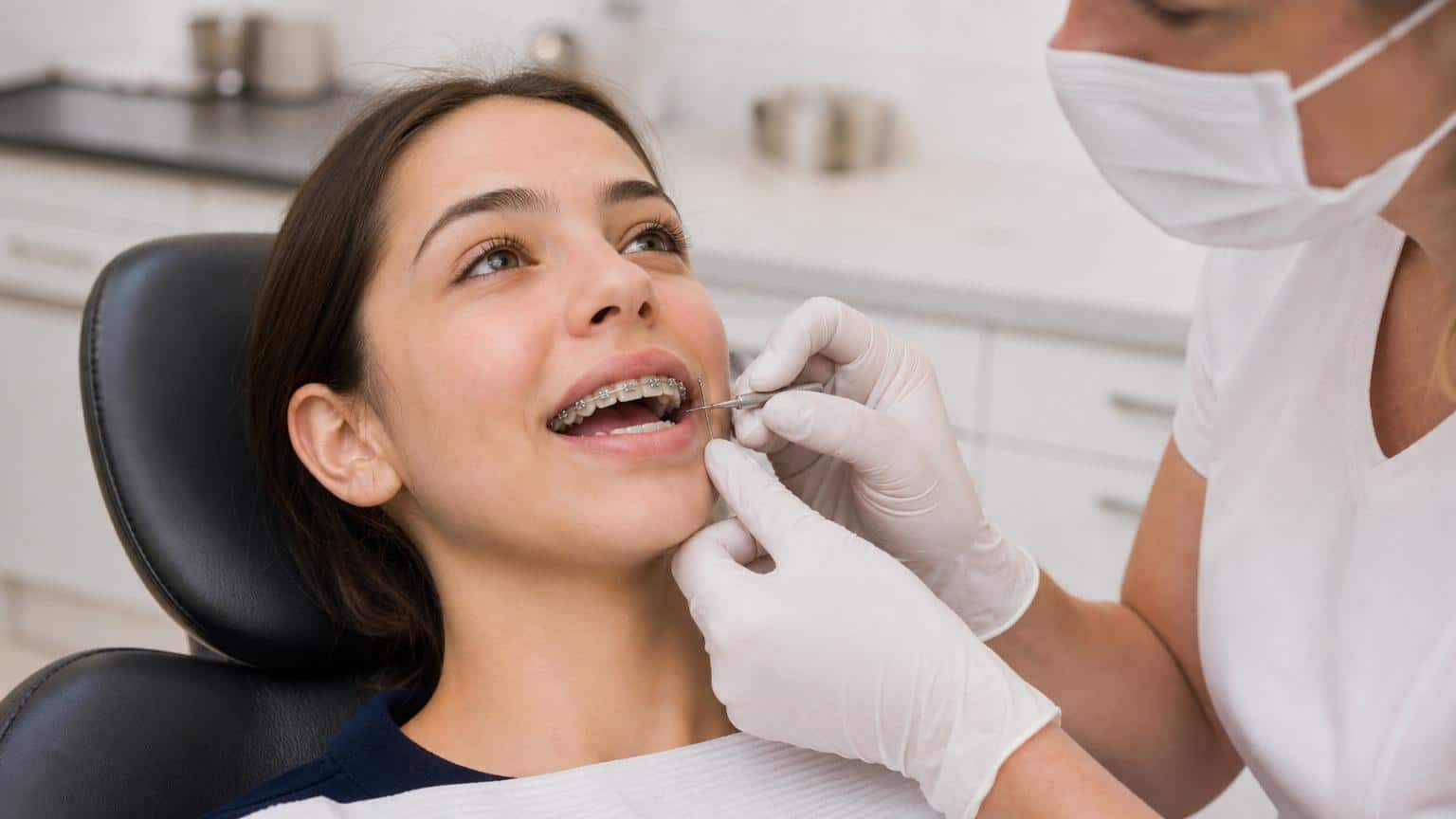 Orthodontist in white gloves and mask using a dental tool to adjust a young woman's metal braces during a clinical appointment.