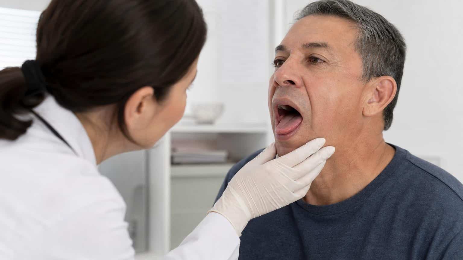 Female doctor in a white coat and gloves examines a middle-aged man's open mouth and throat during a medical checkup.